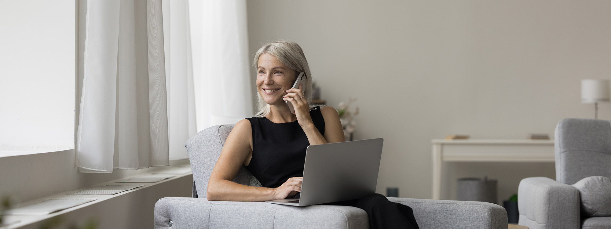 mature female sitting on couch talking on phone with laptop looking out the window and smiling